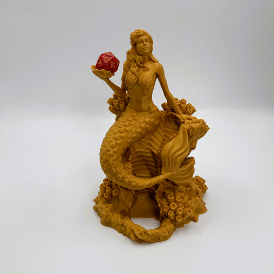 A golden-yellow mermaid figurine sitting on a coral reef base, holding a red gem in one hand, photographed against a neutral light gray background.